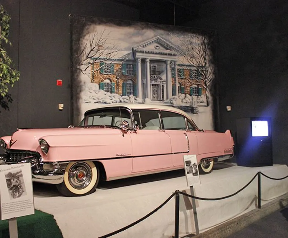 A vintage pink Cadillac is displayed in a museum setting with a large mural of a snowy mansion in the background