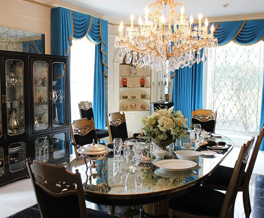 The image shows an opulent dining room with a large ornate chandelier above an oval table set for a meal surrounded by elegant chairs against a backdrop of striking blue draperies and a china cabinet filled with decorative items