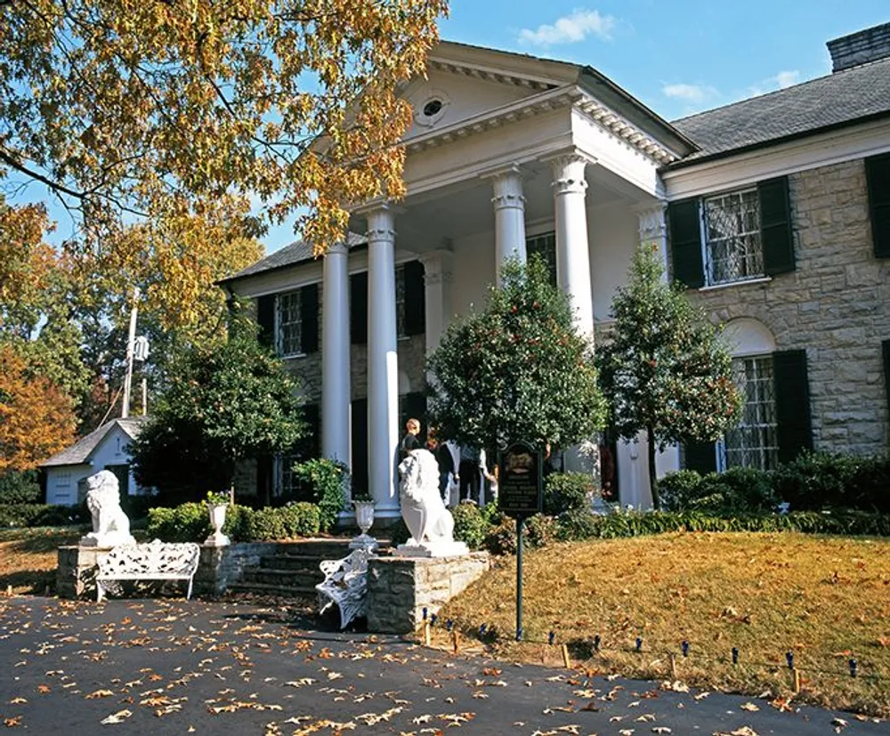 The image shows a stately white-columned building with a person walking in front stone lions at the entrance and a historical marker on the lawn under a sky with scattered clouds