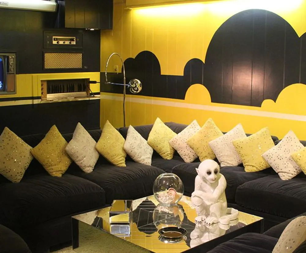 The image shows a retro-styled living room with a striking yellow and black wall design a black sectional sofa adorned with yellow pillows and featuring a vintage television and quirky white owl figurine