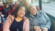 Two smiling women are posing for a photo while sitting on a bus with other passengers in the background.