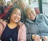 Two women are smiling and sitting together on a bus with other passengers visible in the background