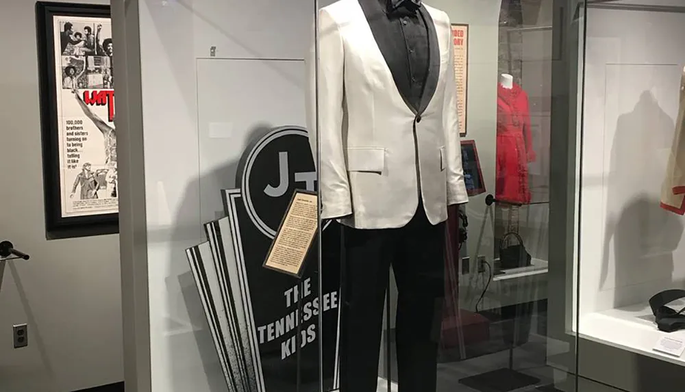 The image shows a museum display featuring a black and white tuxedo with interpretive signs and other exhibits in the background suggesting a retrospective or historical theme