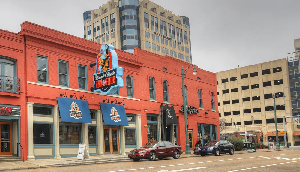 The image shows a vibrant red building featuring the Memphis Music Hall of Fame with a large guitar sign flanked by contemporary buildings under an overcast sky