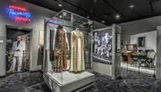 This image shows a music-themed museum exhibit, featuring vintage recording equipment, several iconic stage costumes on display, and large photographs of musicians, under a neon sign reading