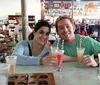 Two people are smiling at the camera while sitting at a table with drinks in a casual indoor setting that looks like a gift shop or cafe