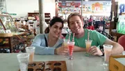 Two people are smiling at the camera while sitting at a table with drinks in a casual indoor setting that looks like a gift shop or cafe.
