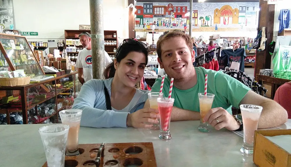 Two people are smiling at the camera while sitting at a table with drinks in a casual indoor setting that looks like a gift shop or cafe