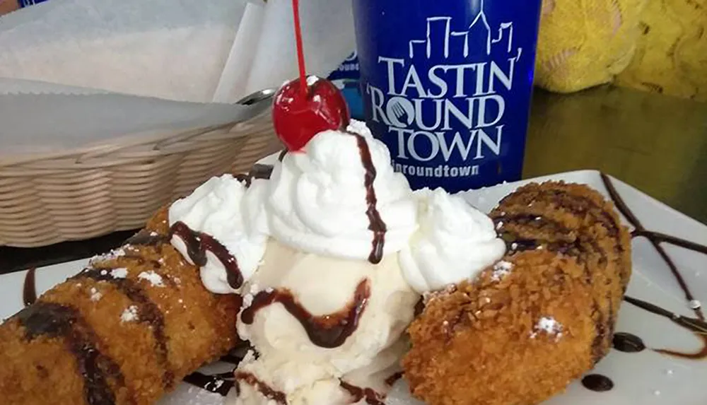 The image shows a decadent dessert featuring deep-fried ice cream topped with whipped cream and a cherry drizzled with chocolate syrup accompanied by a branded blue cup with a logo that reads TASTIN ROUND TOWN