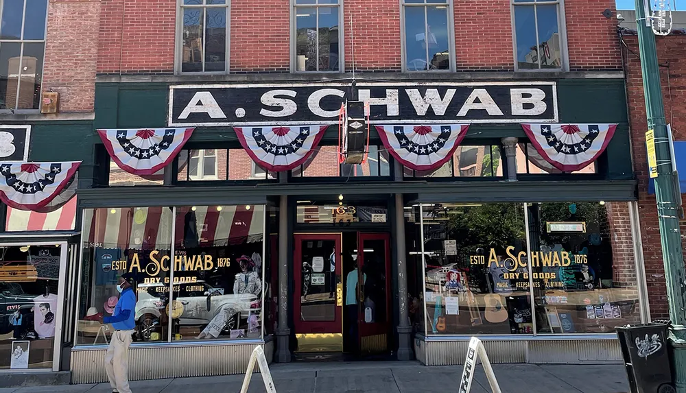 The image shows a person walking in front of the A Schwab store which is adorned with American flag bunting and displays various goods in its windows