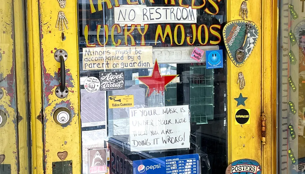 The image shows the entrance to a shop with various eclectic stickers and signs one of which instructs that a mask must cover the nose or its being worn incorrectly