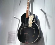 An acoustic guitar with a vintage appearance is displayed on a wall mount, featuring a distinctive pickguard and f-holes.