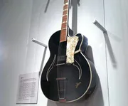 An acoustic guitar with a vintage appearance is displayed on a wall mount, featuring a distinctive pickguard and f-holes.