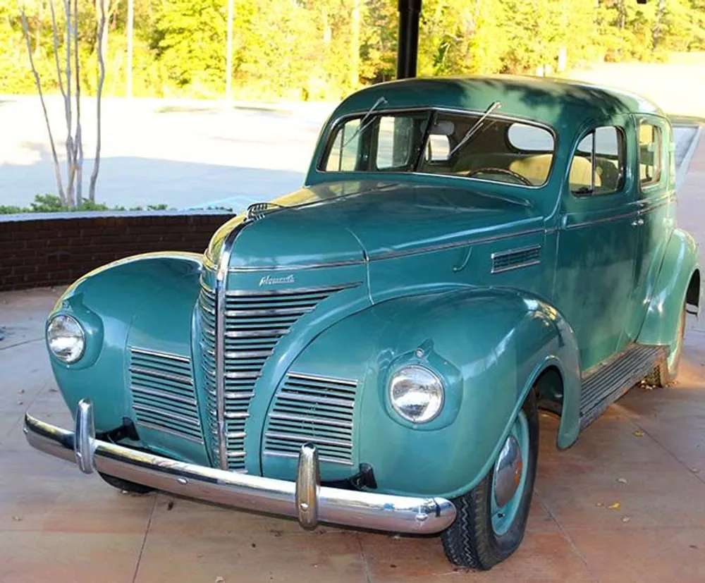 A vintage teal-colored Plymouth automobile is parked under a shelter with trees and daylight in the background