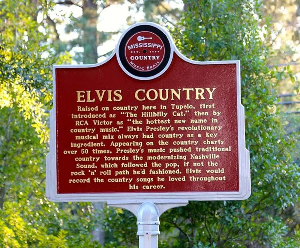 The image shows a historical marker denoting an area called Elvis Country highlighting Elvis Presleys musical roots in country music and his influence on the genre situated in a leafy environment