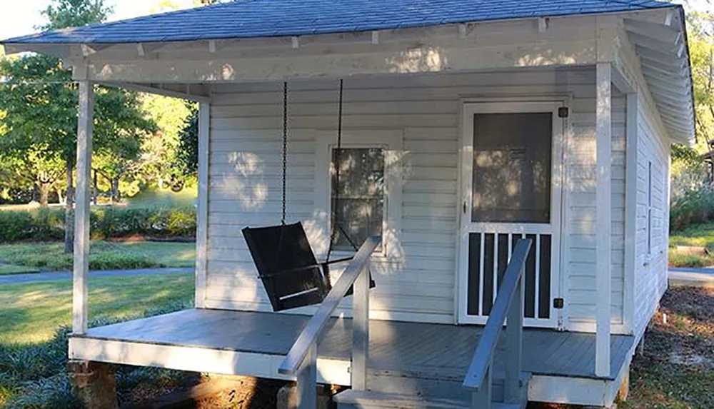 A quaint white cabin with a porch swing basks in the sunlight surrounded by greenery