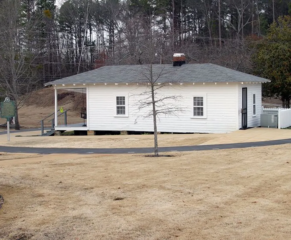 A small white single-story building stands surrounded by a dry grassy area with a historical marker nearby and trees in the background