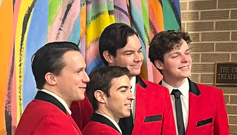 Four men dressed in red jackets and black bow ties are smiling against a colorful backdrop