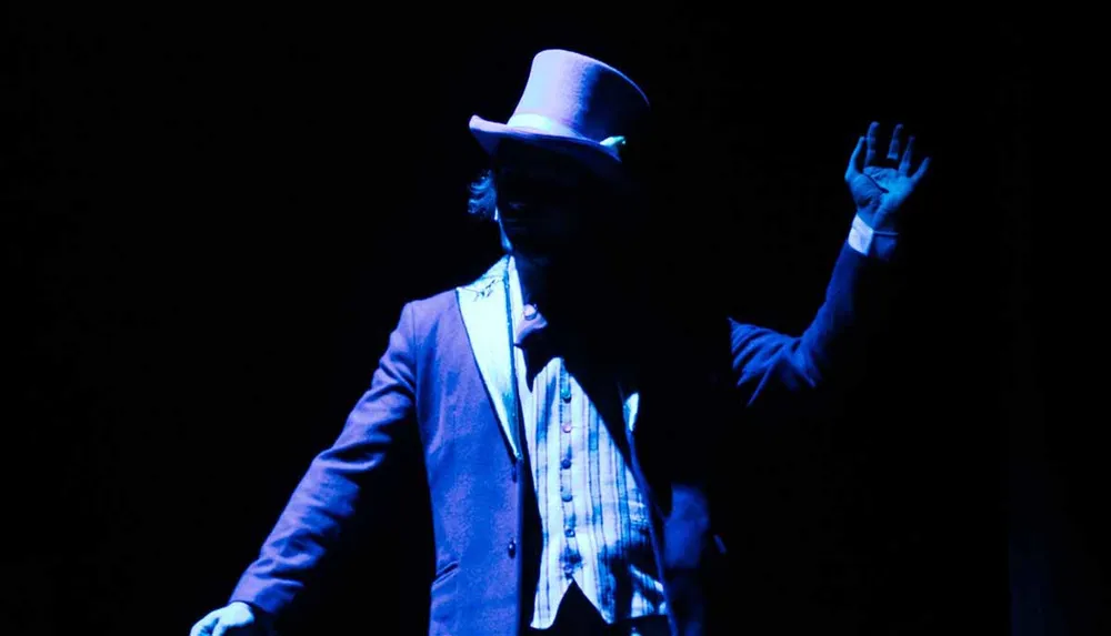 A person wearing a hat and suit is illuminated by blue light against a dark background creating a dramatic silhouette