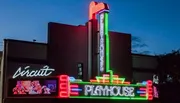 The image shows a brightly lit playhouse with colorful neon signs against a dark evening sky.