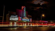 A brightly lit vintage theater marquee stands out against the night sky with streaks of light from passing vehicles blurring in the foreground.