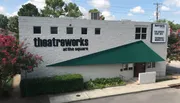 The image shows the exterior of Theatreworks at the Square, with a green awning above the entrance and a sign for current show information, surrounded by greenery and flowering bushes.