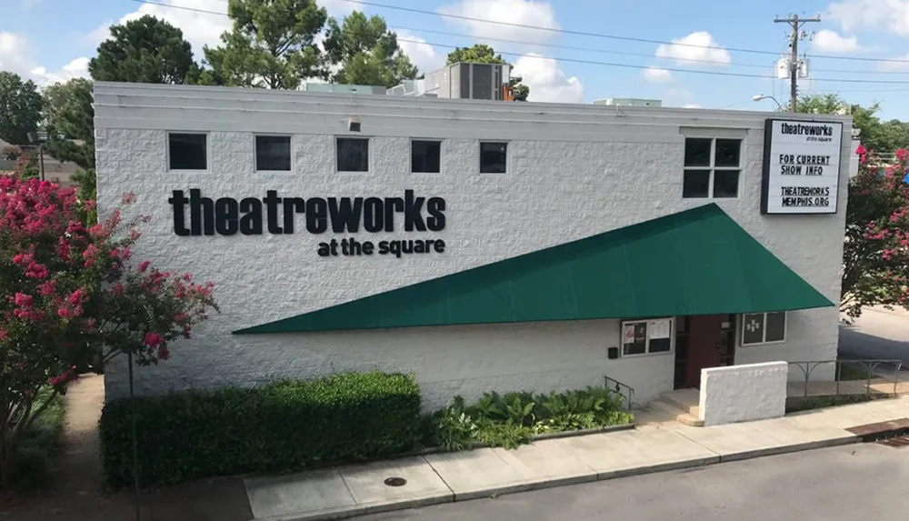 The image shows the exterior of Theatreworks at the Square with a green awning above the entrance and a sign for current show information surrounded by greenery and flowering bushes
