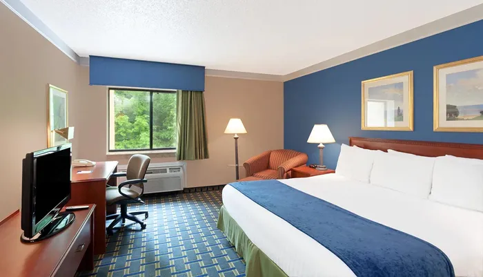 The image shows a neatly arranged hotel room with a bed work desk sitting area and a window overlooking greenery