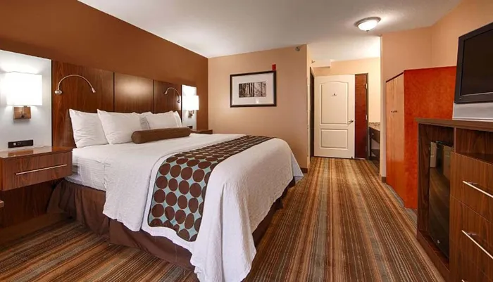 The image shows a neatly arranged hotel room with a large bed wall-mounted bedside lights a TV and a small kitchenette area visible in the background