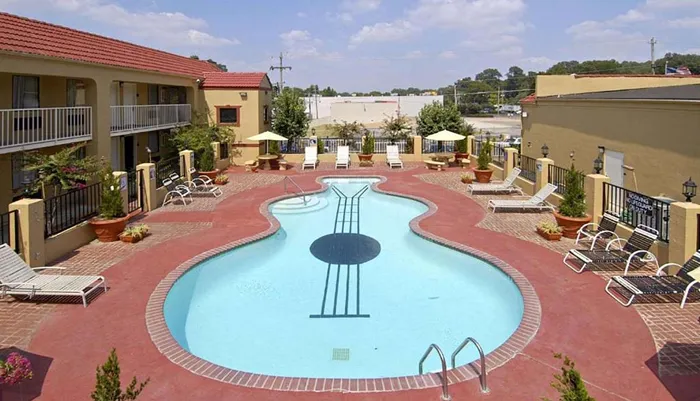 The image features a guitar-shaped swimming pool surrounded by lounge chairs with an adjacent building featuring balconies indicating a hotel or apartment setting
