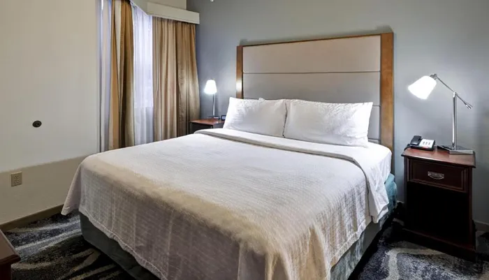 This is a neatly presented hotel room with a large bed two bedside tables with lamps and a drawn curtain in the background