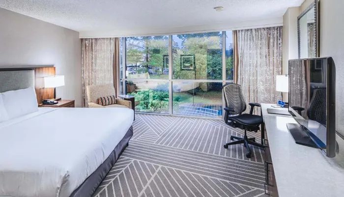 The image shows a neatly arranged hotel room with a large bed a work desk with a chair a television and a full-length window offering a view of trees outside