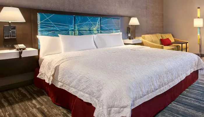 The image shows a neatly made king-sized bed with white linens in a well-furnished hotel room featuring modern dcor and a cozy sitting area