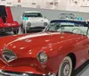 The image shows a collection of classic cars with a prominent red convertible in the foreground inside a museum or exhibition setting