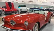 The image shows a collection of classic cars with a prominent red convertible in the foreground inside a museum or exhibition setting.