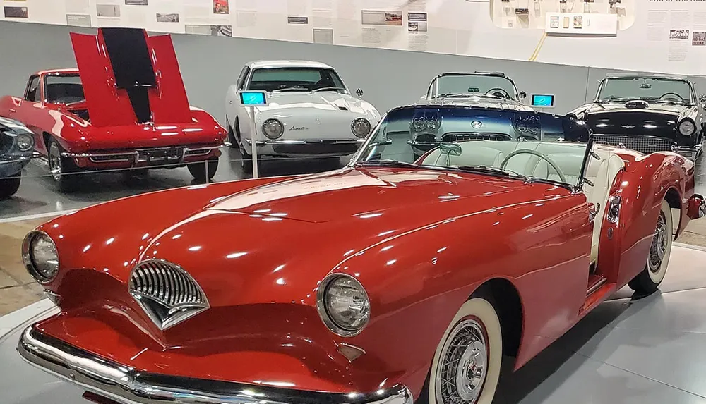 The image shows a collection of classic cars with a prominent red convertible in the foreground inside a museum or exhibition setting