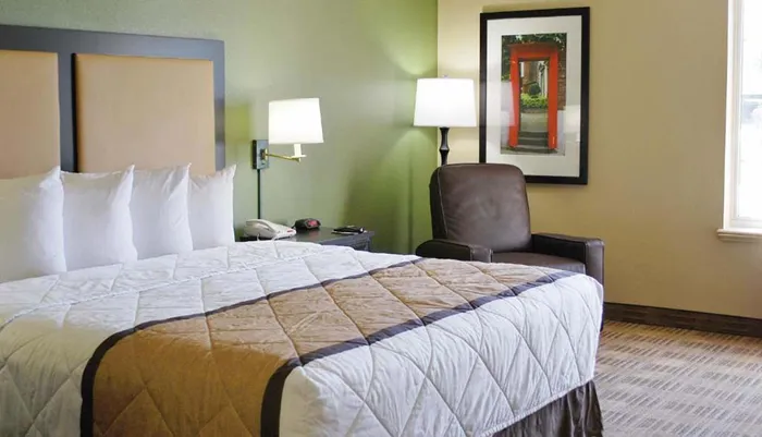 The image shows a neatly arranged hotel room with a large bed side lamps artwork on the wall and a comfortable chair creating a cozy atmosphere