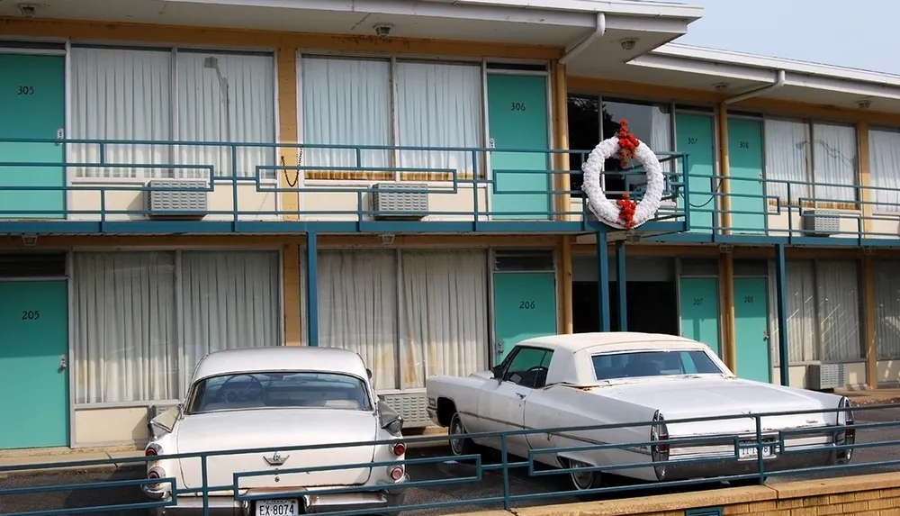 The image shows a two-story vintage motel with a wreath on the railing and classic cars parked in front of the rooms