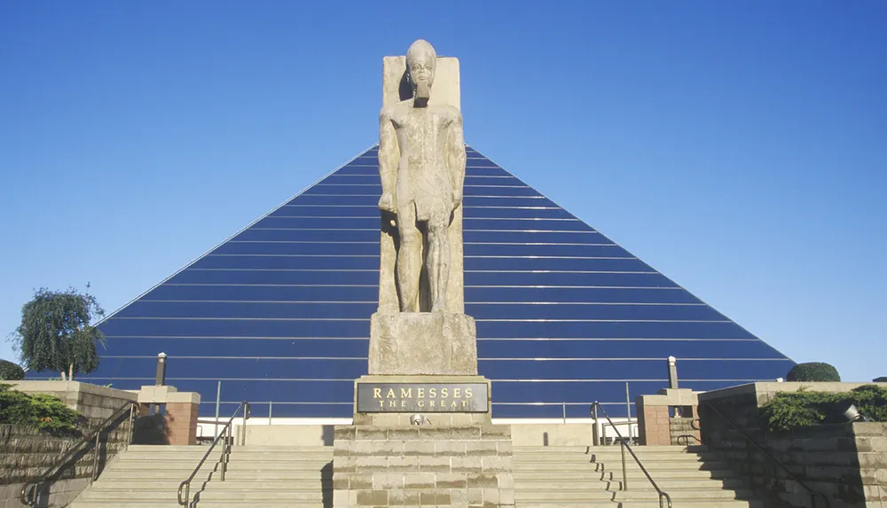 The image shows a statue of Ramesses the Great in front of a modern building with a pyramidal glass facade under a clear blue sky