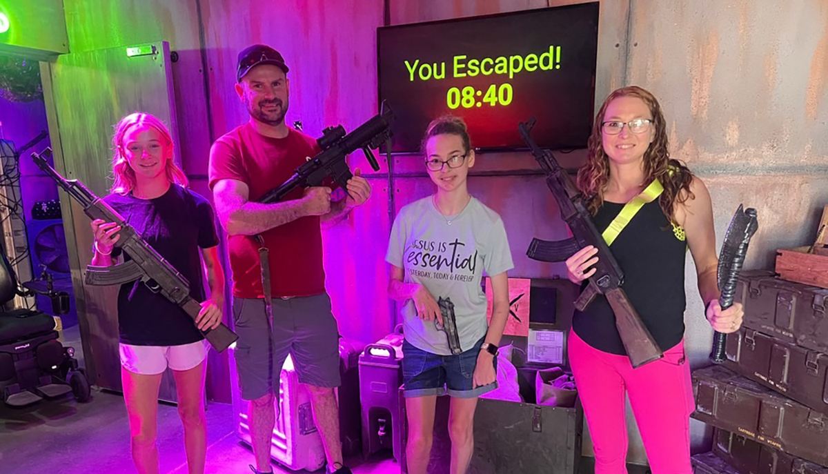 A group of four people is standing in an escape room setting, holding replica guns, with a screen behind them displaying 