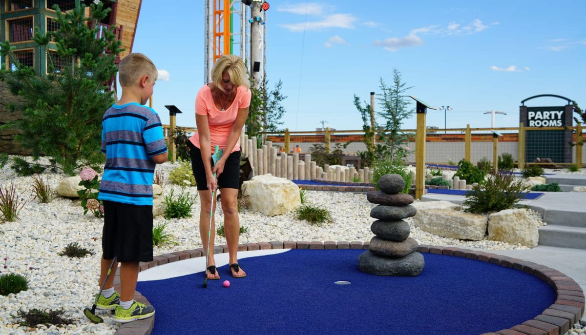 A woman is playing mini-golf with a boy on a course with blue artificial turf, surrounded by decorative rocks and plants.