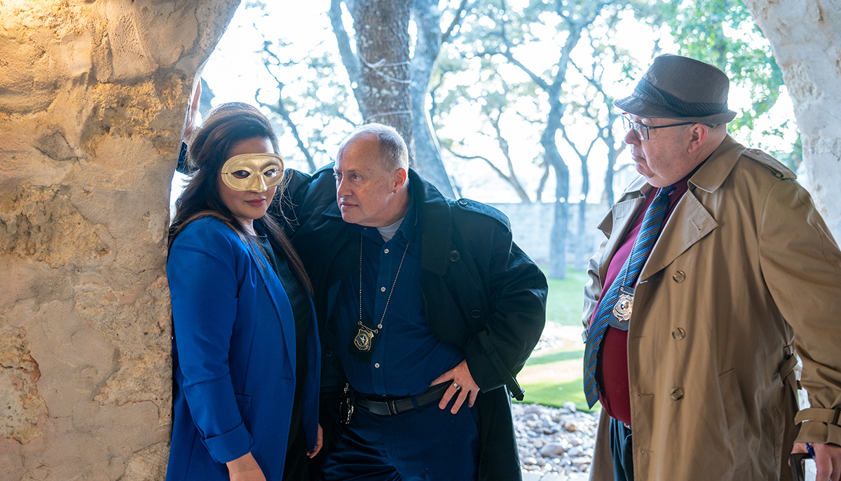 Two detectives are questioning a woman in a mask standing against a stone wall.