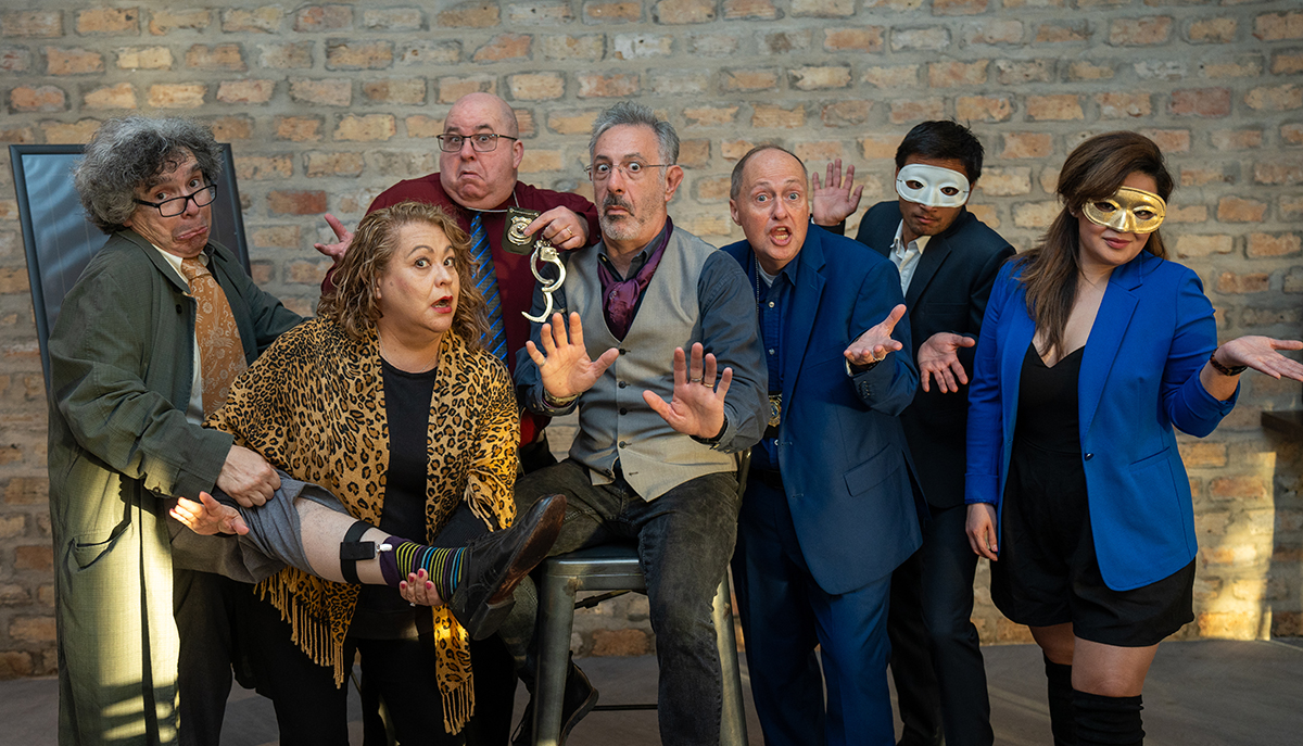 A group of seven people, some wearing masks or holding handcuffs, pose humorously in front of a brick wall, with one person sitting and others gesturing dramatically.