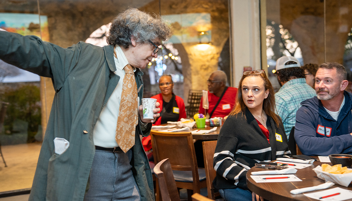 A man in a coat is gesturing while holding a cup and engaging with people seated at a round table during a social gathering.
