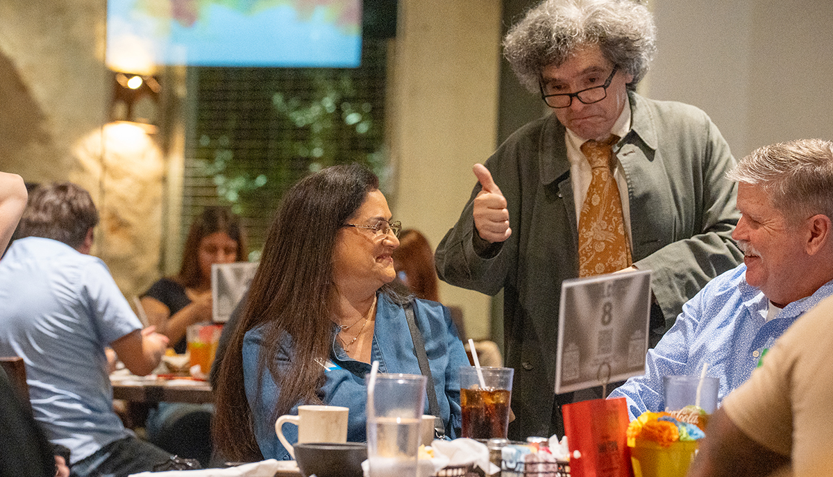 A man with curly hair and glasses gives a thumbs-up to a woman and a man sitting at a table at a casual dining restaurant.