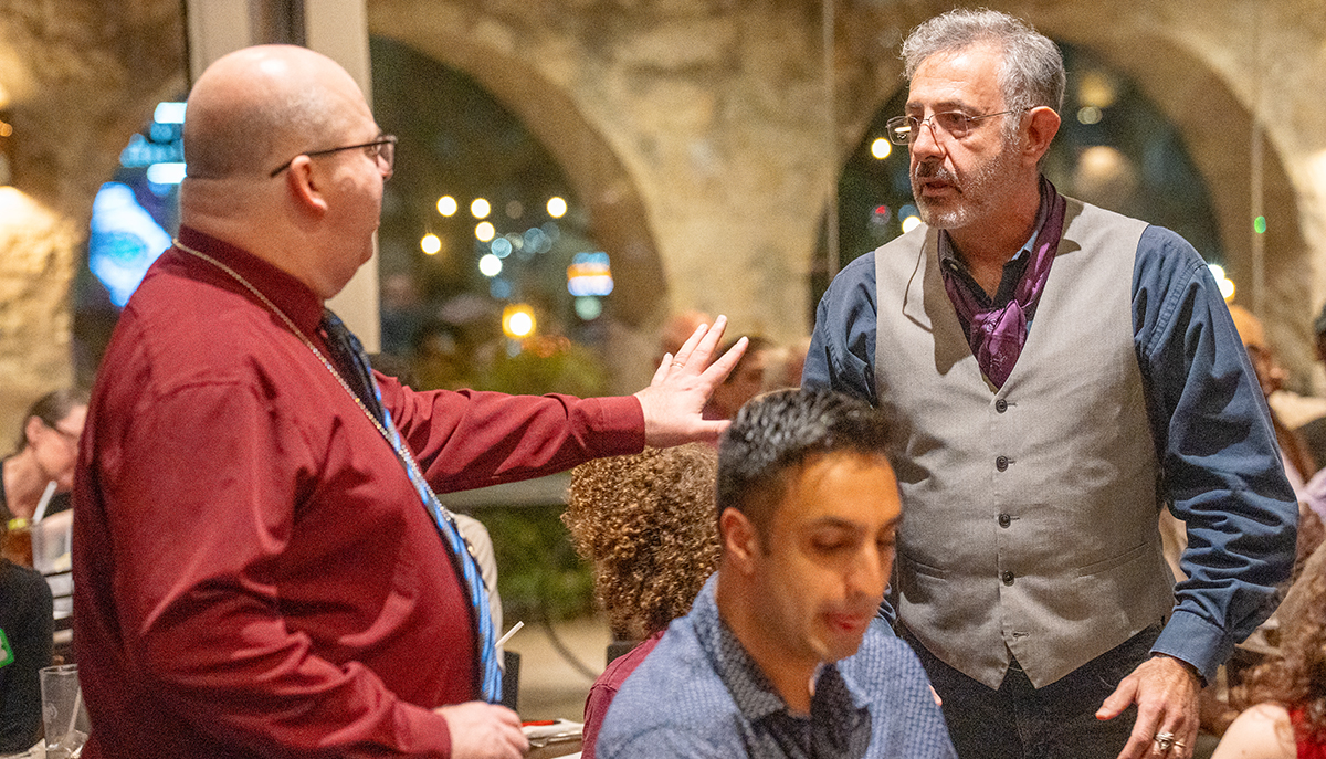 Two men are having a conversation in a lively restaurant setting, with blurred patrons and lights in the background.