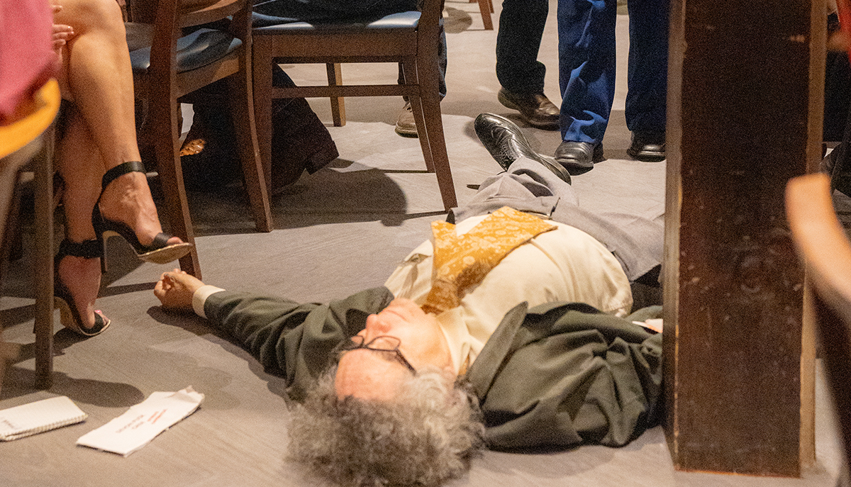 A person is lying on the floor in a crowded room, with people standing and sitting nearby.