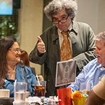 A person with curly hair wearing a coat gives a thumbs-up to two seated individuals at a dining table.