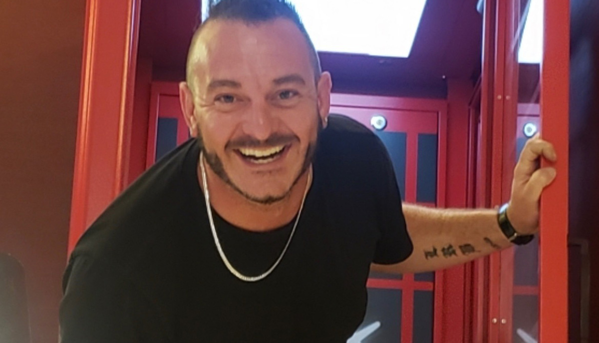 A smiling man with a beard is standing in a brightly lit room, holding onto a red doorframe.