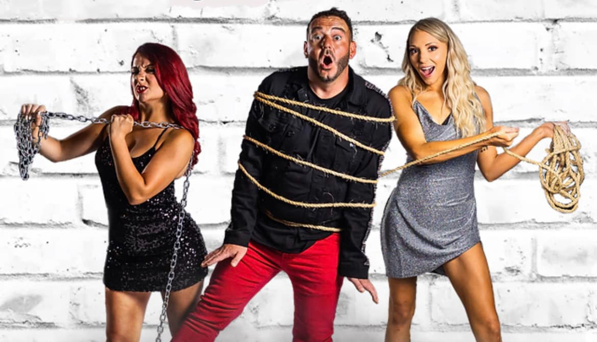 Three people are standing in front of a white brick wall, with the man in the middle tied up in rope and women on either side holding chains and rope, looking surprised and amused.